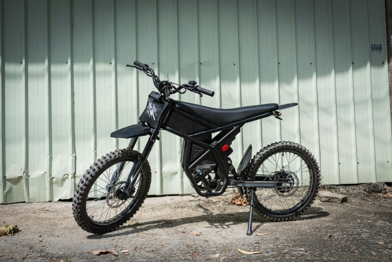 A modern black electric dirt bike parked against a corrugated metal wall on a sunny day.